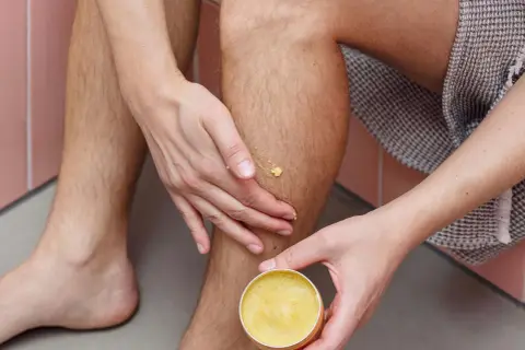 Hände tragen eine gelbe Salbe auf ein Bein auf. Manche glauben, sheabutter trocknet haut aus, aber diese Salbe spendet Feuchtigkeit.