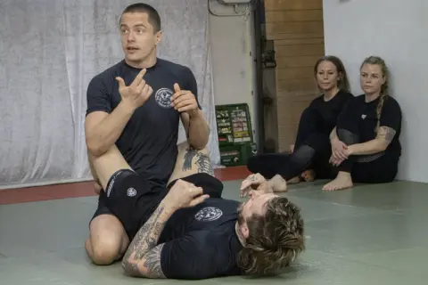 Instruktör visar en teknik i stark bjj. Två kvinnor sitter och observerar.