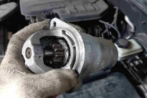 Defekter Anlasser Mercedes: Werkzeug in Handschuh hält ausgebauten Anlasser mit sichtbarem Ritzel. Kosten für Reparatur im Blick.