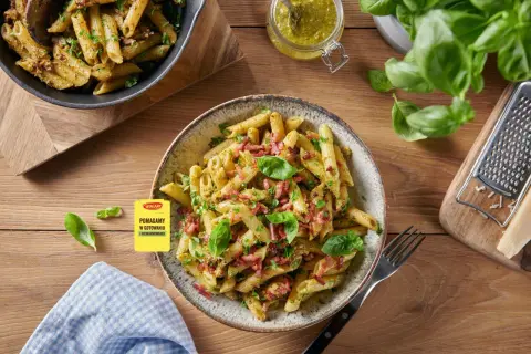 Makaron penne z pesto, boczkiem i bazylią na talerzu, obok słoiczek z pesto i tarką.
