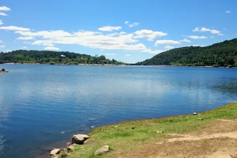 Un día soleado en la ruta del agua La Jarosa, con un embalse azul rodeado de colinas verdes y un cielo con nubes blancas.