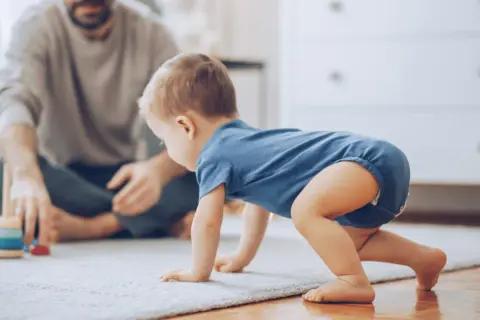 Dziecko w niebieskim body raczkuje na dywanie, obok siedzi ojciec z zabawkami.