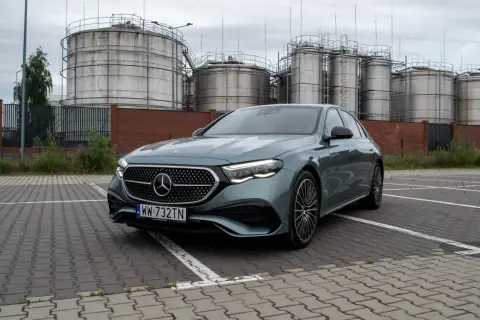 Nowy Mercedes E220d w kolorze zielonym, zaparkowany przed przemysłowymi zbiornikami.