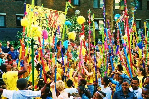 Gente celebra en un desfile, levantando coloridos bastones adornados con pompones y cintas. Un cartel dice "POLE-O-NG STATION". Parece una fiesta, quizás un bank holiday.