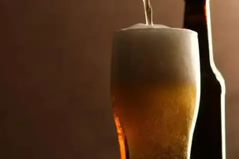 Birra pils che si versa in un bicchiere, con una bottiglia sullo sfondo. Schiuma abbondante e colore dorato invitante.