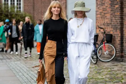 Deux femmes marchent, l'une porte un pantalon bicolore audacieux, l'autre un ensemble blanc décontracté.