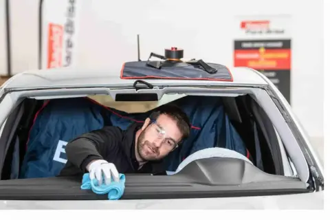 Un technicien porte des lunettes de protection et utilise un chiffon bleu pour nettoyer l'intérieur du pare-brise d'une voiture.