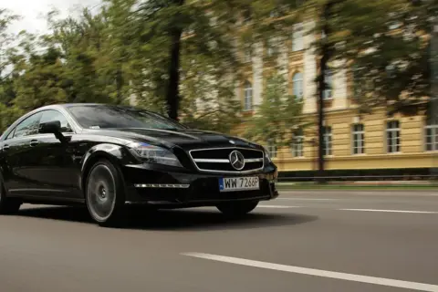 Czarny Mercedes CLS 63 AMG Coupe pędzi ulicą, mijając zabytkową kamienicę i drzewa.