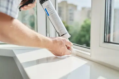 Mężczyzna aplikuje masę uszczelniającą, pokazując jak zamontować parapet wewnętrzny. Biały parapet i okno.