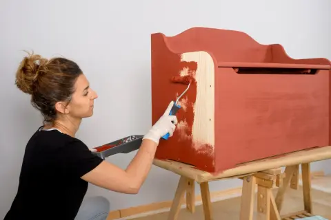 Mujer aplica pintura para aglomerado a un baúl de madera con un rodillo.