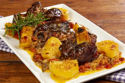 Un saporito agnello con patate, cotto lentamente con pomodorini e rosmarino, servito su un piatto bianco.