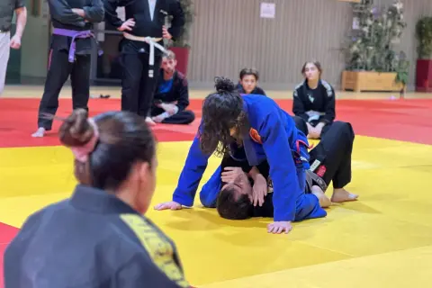 Deux pratiquants de jiu jitsu s'entraînent sur un tapis jaune et rouge. L'un est en position dominante, l'autre en défense.