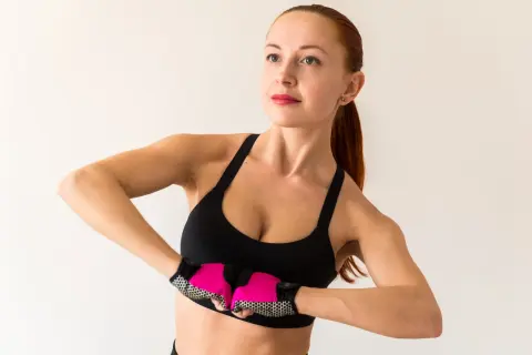 Kobieta w sportowym topie wykonuje ćwiczenia na podniesienie biustu, z różowymi rękawiczkami na dłoniach.