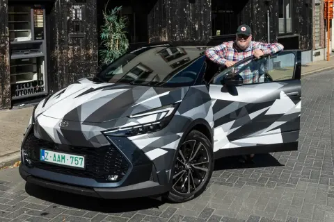 Man lutar sig mot en Toyota C-HR Hybrid i kamouflagemönster. Bilen är parkerad på en kullerstensgata framför en byggnad med en automat.