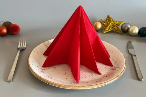 Un sapin en serviette papier rouge décore une assiette, prêt pour un repas festif. Des boules de Noël et une étoile dorée complètent la mise en scène.