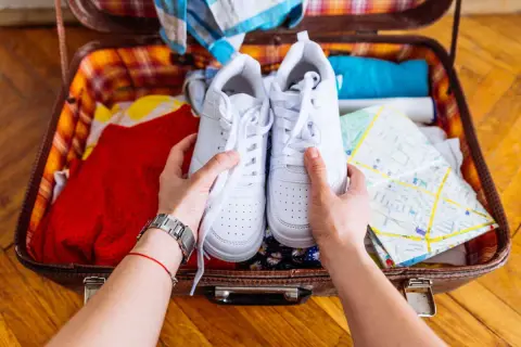 Comment ranger des chaussures dans une valise : une paire de baskets blanches est tenue au-dessus d'une valise ouverte remplie de vêtements et d'une carte.