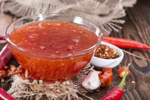 En skål med söt chili sås omgiven av färska och torkade chilifrukter, vitlök och pepparkorn.