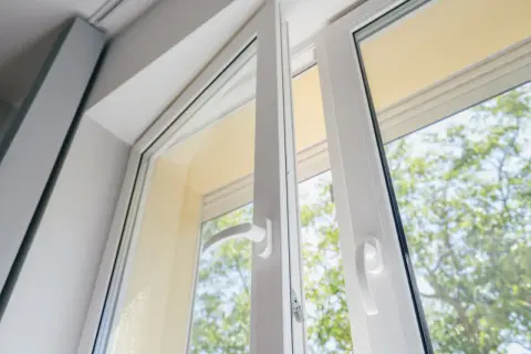 Białe okno z profili pvc, uchylone, z widokiem na zielone drzewa.