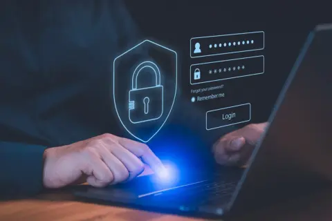 Dłoń dotyka ekranu laptopa z interfejsem logowania. Ikona tarczy z kłódką symbolizuje bezpieczeństwo, a obok pola na login i hasło znajduje się przycisk "Login". To nowoczesny menedżer haseł.
