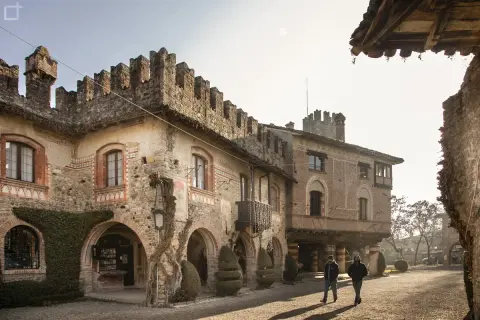 Antico borgo Visconti a Piacenza, con torri merlate e archi in pietra. Due uomini passeggiano nel cortile illuminato dal sole.