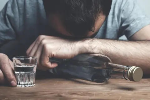 Zrezygnowany mężczyzna opiera głowę na butelce alkoholu, obok stoi kieliszek. Czy alkohol podnosi cholesterol?