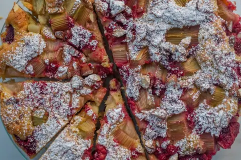 Pyszne ciasto jogurtowe z rabarbarem i malinami, posypane cukrem pudrem. Idealne na letnie popołudnie.