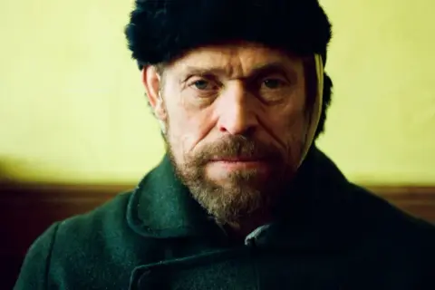 Willem Dafoe w czapce z futra i ciemnym płaszczu, w kadrze z filmu.