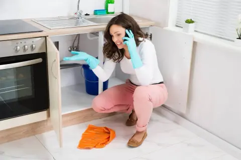 Une femme en gants bleus, au téléphone, tente de colmater une fuite d'eau sous son évier, un seau bleu à ses pieds.