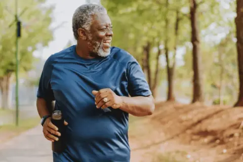 Un homme souriant court dans un parc, tenant une bouteille d'eau. Il est un exemple d'exercices pour perdre du ventre après 60 ans.