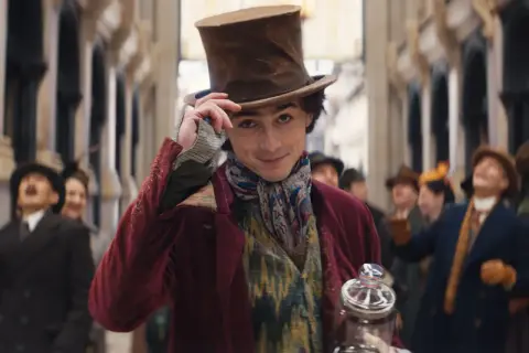 Timothée Chalamet w filmach, jako młody Willy Wonka, w cylindrze i barwnym stroju, z uśmiechem pozdrawia tłum.