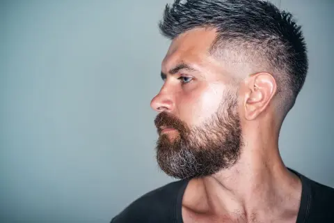 Mann mit Bart und tiefer fasson haarschnitt, im Profil auf grauem Hintergrund.