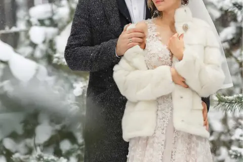 Para młoda w zimowej scenerii. Ona w białym futerku i koronkowej sukni, on w eleganckim płaszczu. Ich ślub w zimie wygląda bajkowo.