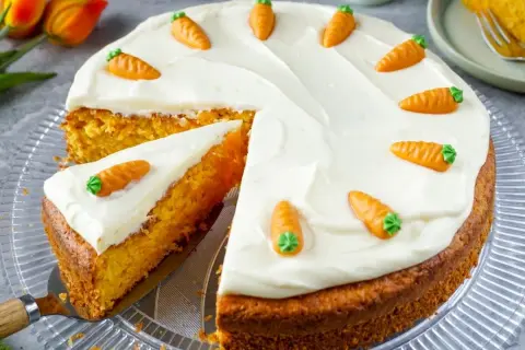 Saftiger Karottenkuchen mit cremiger Glasur und kleinen Marzipan-Karotten. Ein Stück ist bereits angeschnitten.
