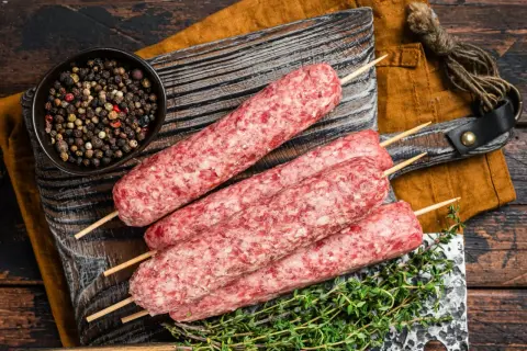 Surowe kofty wołowe na patyczkach, gotowe do grillowania, obok miseczki z pieprzem i gałązki tymianku.