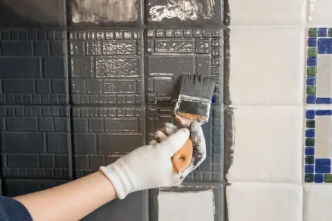 Ręka w białej rękawiczce maluje pędzlem szare płytki w łazience, odświeżając ich wygląd.
