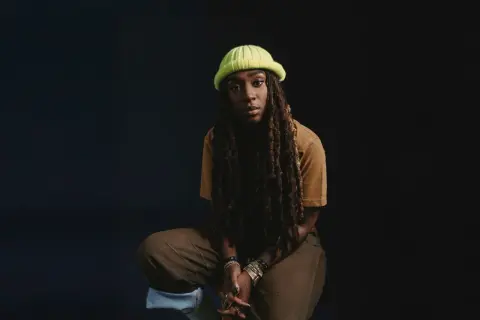 Jackie Hill Perry, mit Dreadlocks und einer neongrünen Mütze, sitzt nachdenklich vor dunklem Hintergrund.