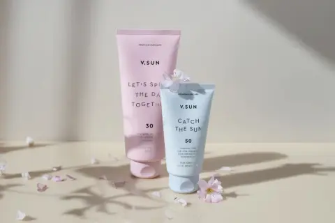 Zwei V.SUN Sonnencreme-Tuben, eine mit LSF 30 für den Körper, die andere mit LSF 50 für das Gesicht, umgeben von zarten Blütenblättern.