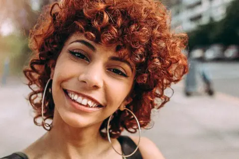 Sonrisa radiante de una mujer con rizos castaño rojizo y grandes aros plateados.