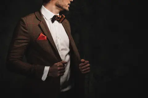 Elegancki mężczyzna w brązowej, tweedowej marynarce w kratkę, białej koszuli i muszce. Stylizacje z tweedową marynarką to klasyka.