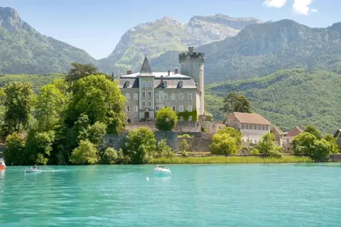 Un château majestueux surplombe un lac turquoise, invitant à aller à Annecy. Des montagnes verdoyantes en arrière-plan.