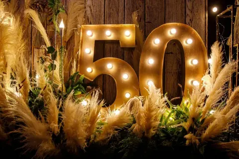Złote litery "50" z żarówkami, otoczone puszystymi trawami i zielenią, tworzą magiczną dekorację na 50 urodziny.