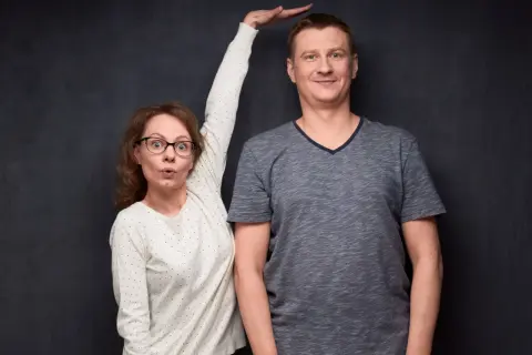 Kobieta z wyrazem zaskoczenia na twarzy mierzy wzrost mężczyzny, przykładając dłoń do jego głowy.