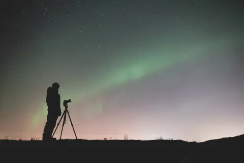 Sylwetka fotografa z aparatem na statywie pod rozgwieżdżonym niebem i zorzą polarną. Efekt bokeh gwiazd dodaje magii.