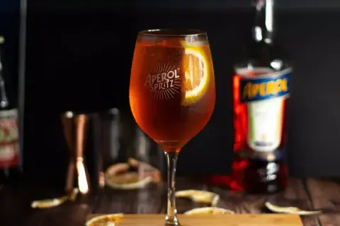 Szklanka Aperol Spritz z plasterkiem pomarańczy, butelka Aperol i inne składniki na drewnianym stole.