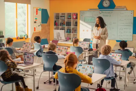 Nauczycielka prowadzi lekcję matematyki w nowoczesnej klasie. Dzieci uczą się z laptopów, co pokazuje postęp w pracy w oświacie.