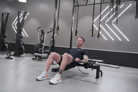Man utför hip thrusters med skivstång på en bänk i ett modernt gym.