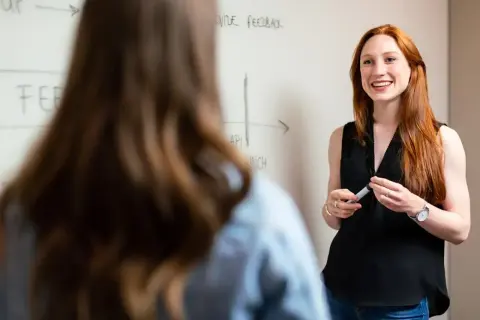 En kvinna med rött hår ler och ger positiv återkoppling vid en whiteboard. Hon håller en penna.