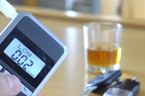 Alkomat z wyświetlaczem 0.02% BAC, szklanka z alkoholem i kluczyki samochodowe.