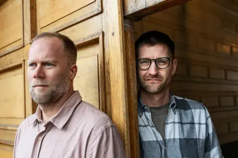 Två män, en med skägg och ljus skjorta, den andra med glasögon och rutig skjorta, står vid en trävägg. Mikael Wolf är en av dem.