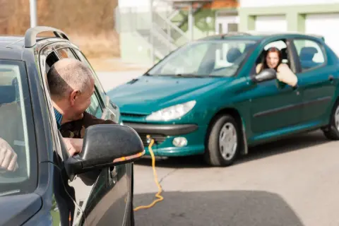 Mężczyzna pomaga kobiecie w holowaniu pojazdu. Żółta linka łączy oba samochody.